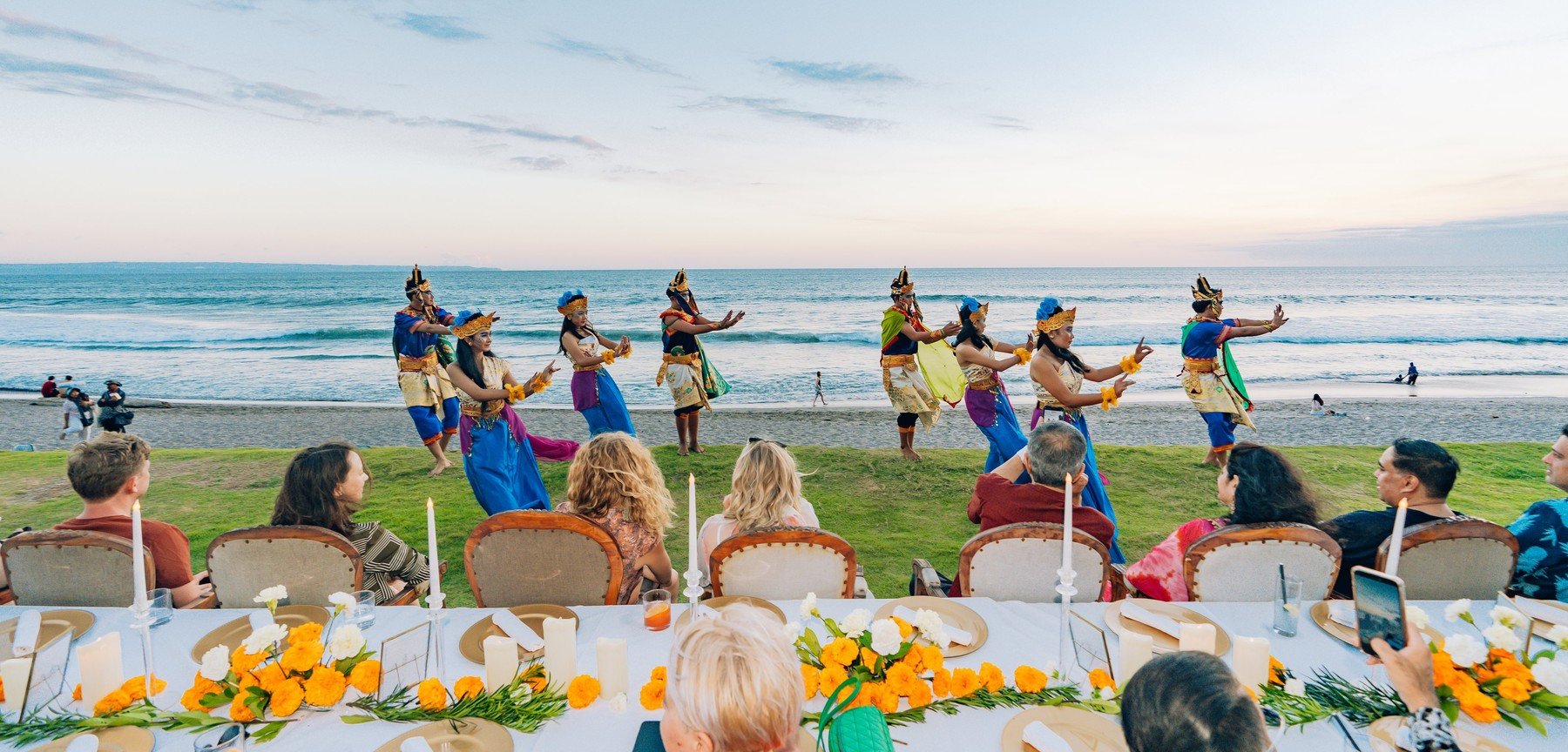 Balinese dancers performing on the lawn above the beach at dusk, guests watching from a long candlelit table dressed with marigolds